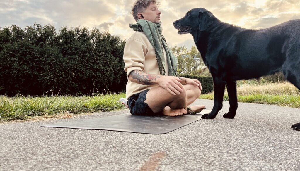 Ein Mann sitzt barfuß im Schneidersitz auf einer Yogamatte auf einem Weg, meditiert mit geschlossenen Augen und gefalteten Händen, während ihn ein schwarzer Labrador aus nächster Nähe interessiert beobachtet, im Hintergrund eine Hecke und der Abendhimmel.