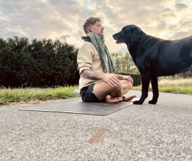 Ein Mann sitzt barfuß im Schneidersitz auf einer Yogamatte auf einem Weg, meditiert mit geschlossenen Augen und gefalteten Händen, während ihn ein schwarzer Labrador aus nächster Nähe interessiert beobachtet, im Hintergrund eine Hecke und der Abendhimmel.