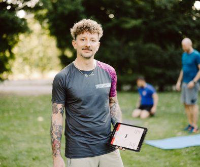Tätowierter Fitnesstrainer hält ein Tablet bei einem Outdoor-Gruppentraining im Park