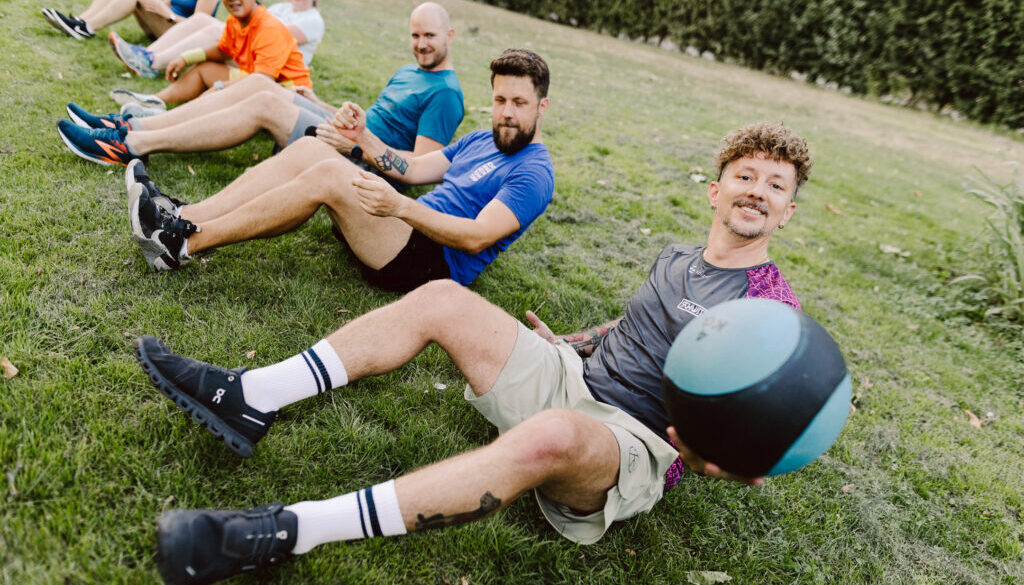 eihe von fünf Personen liegt seitlich versetzt im Sit-up auf einer Wiese und hält sich gegenseitig an den Händen; die vorderste Person im grauen SCHWITZEREI-Shirt hält lächelnd einen türkisen Medizinball in die Kamera.