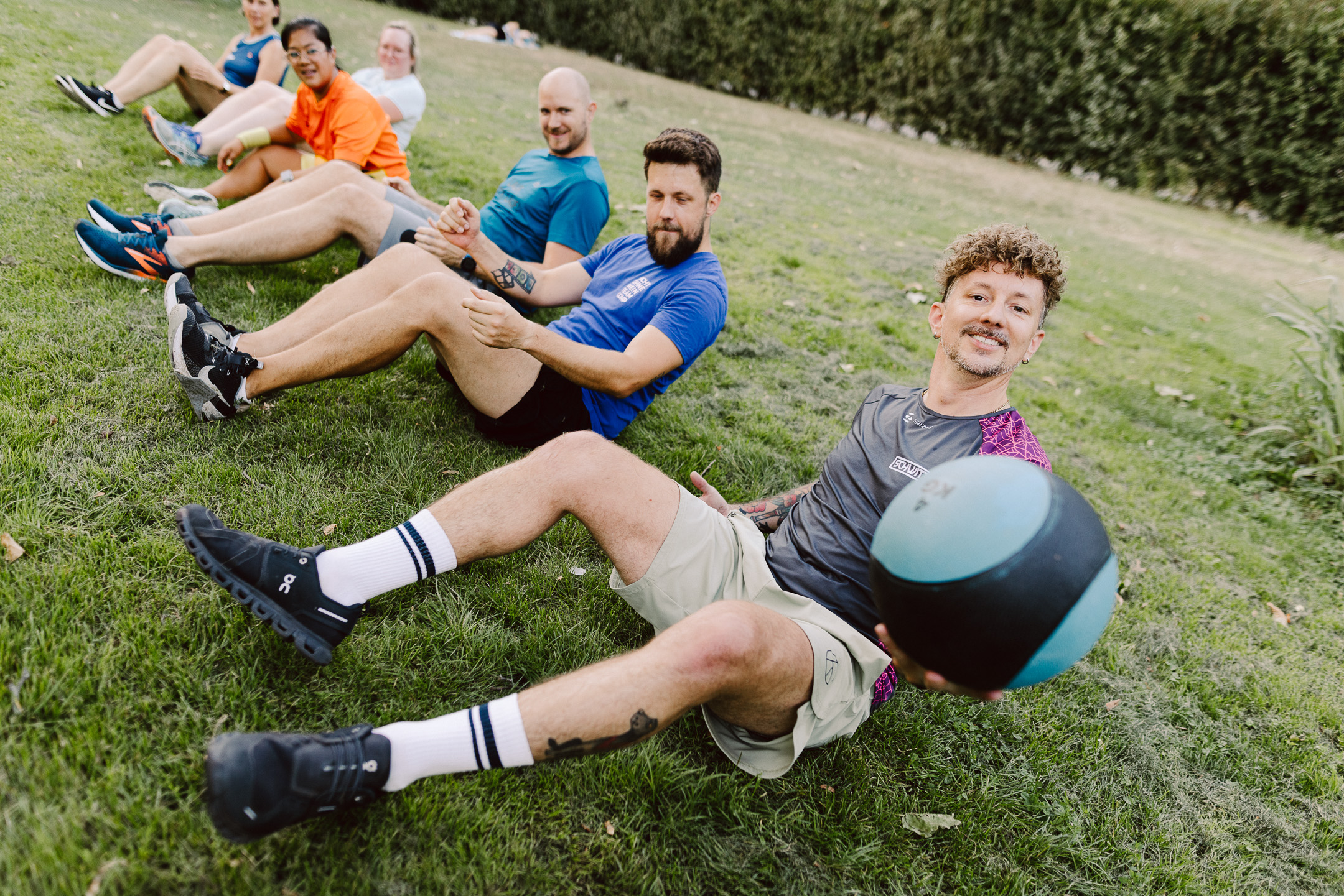 eihe von fünf Personen liegt seitlich versetzt im Sit-up auf einer Wiese und hält sich gegenseitig an den Händen; die vorderste Person im grauen SCHWITZEREI-Shirt hält lächelnd einen türkisen Medizinball in die Kamera.