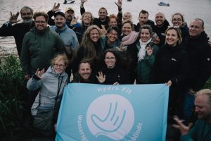 DSC03283 Gruppe am Wasser mit Banner von Hydro Fit – Wassersport für Taubblinde; viele machen Gebärdenzeichen
