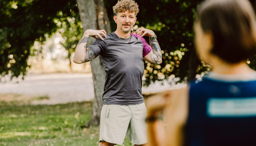 Ein Trainer mit lockigen Haaren, Tattoos und grauem SCHWITZEREI-Shirt steht im Park und führt eine Schultermobilisationsübung vor, während zwei Teilnehmerinnen im Vordergrund unscharf zusehen.
