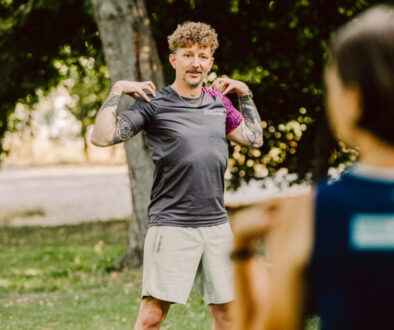 Ein Trainer mit lockigen Haaren, Tattoos und grauem SCHWITZEREI-Shirt steht im Park und führt eine Schultermobilisationsübung vor, während zwei Teilnehmerinnen im Vordergrund unscharf zusehen.
