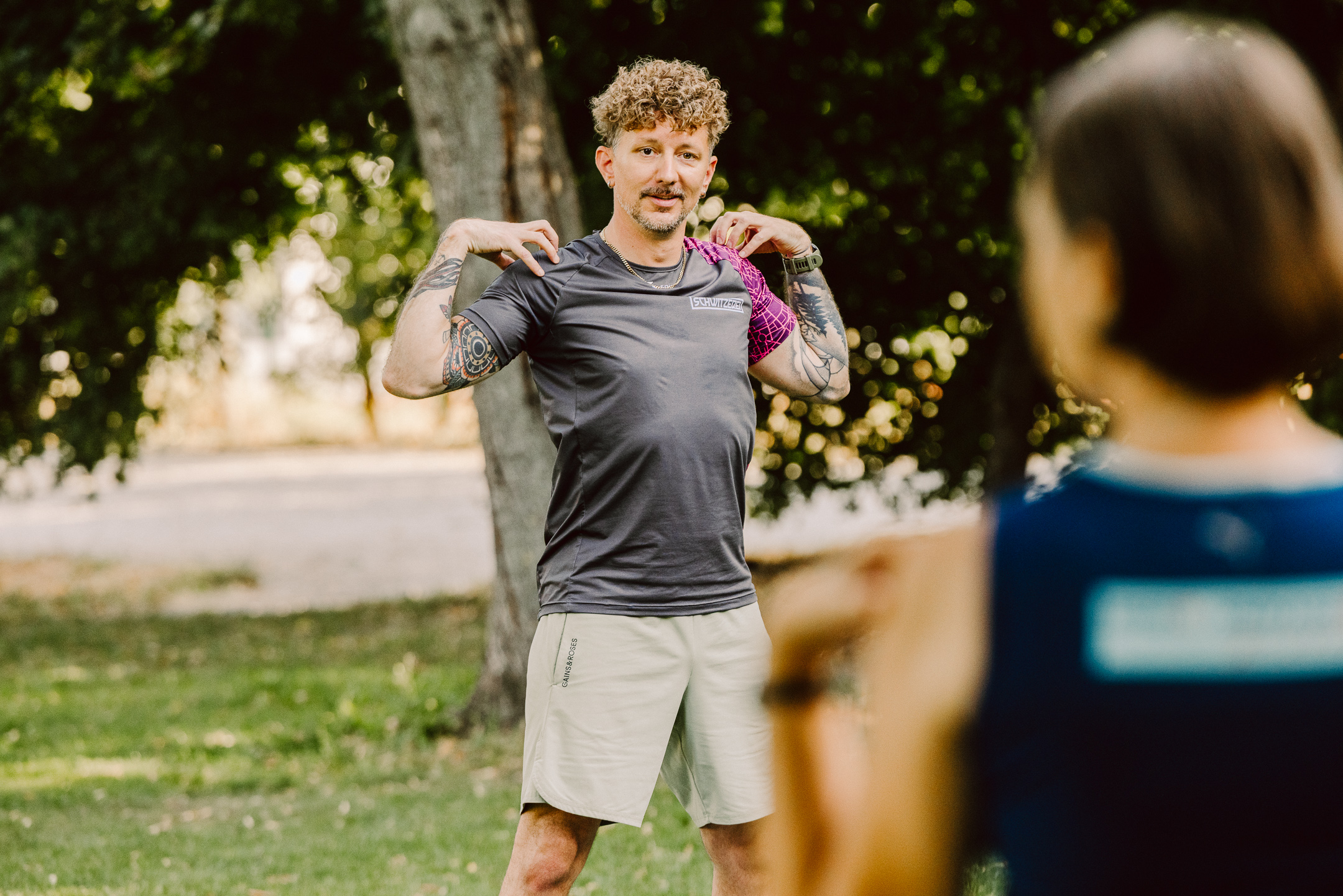 Ein Trainer mit lockigen Haaren, Tattoos und grauem SCHWITZEREI-Shirt steht im Park und führt eine Schultermobilisationsübung vor, während zwei Teilnehmerinnen im Vordergrund unscharf zusehen.