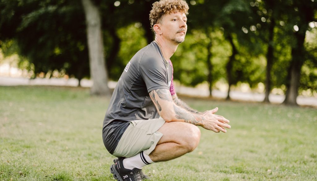 Ein Mann mit lockigem Haar und Tätowierungen hockt in Sportkleidung auf einer Wiese vor Bäumen