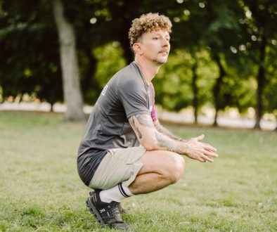 Ein Mann mit lockigem Haar und Tätowierungen hockt in Sportkleidung auf einer Wiese vor Bäumen