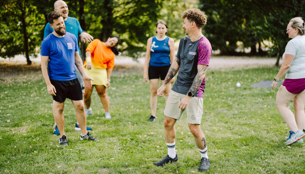 Trainer lacht gemeinsam mit sechs Teilnehmenden während eines Outdoor-Gruppentrainings auf einer Wiese.