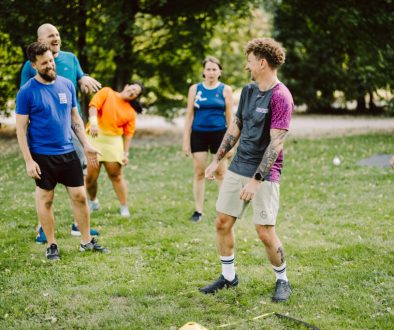 Trainer lacht gemeinsam mit sechs Teilnehmenden während eines Outdoor-Gruppentrainings auf einer Wiese.