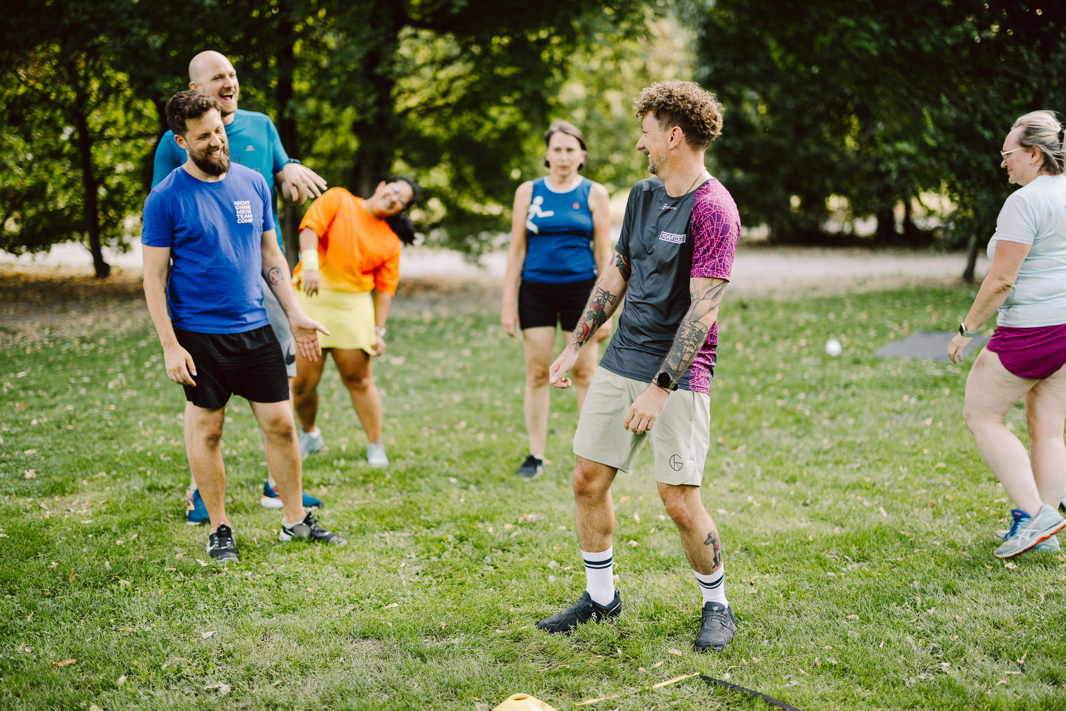Trainer lacht gemeinsam mit sechs Teilnehmenden während eines Outdoor-Gruppentrainings auf einer Wiese.