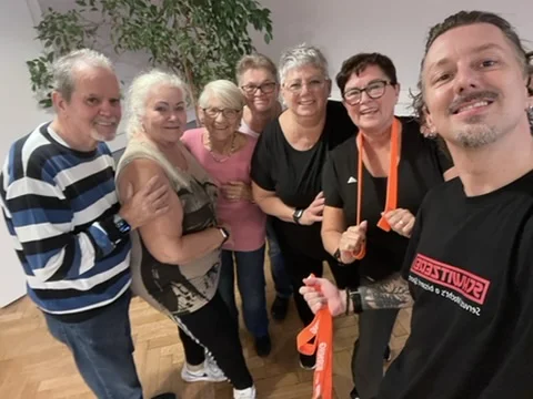 Selfie in einem Raum: Trainer Manfred und sieben Teilnehmende lächeln in die Kamera. Einige halten orange Trainingsbänder.