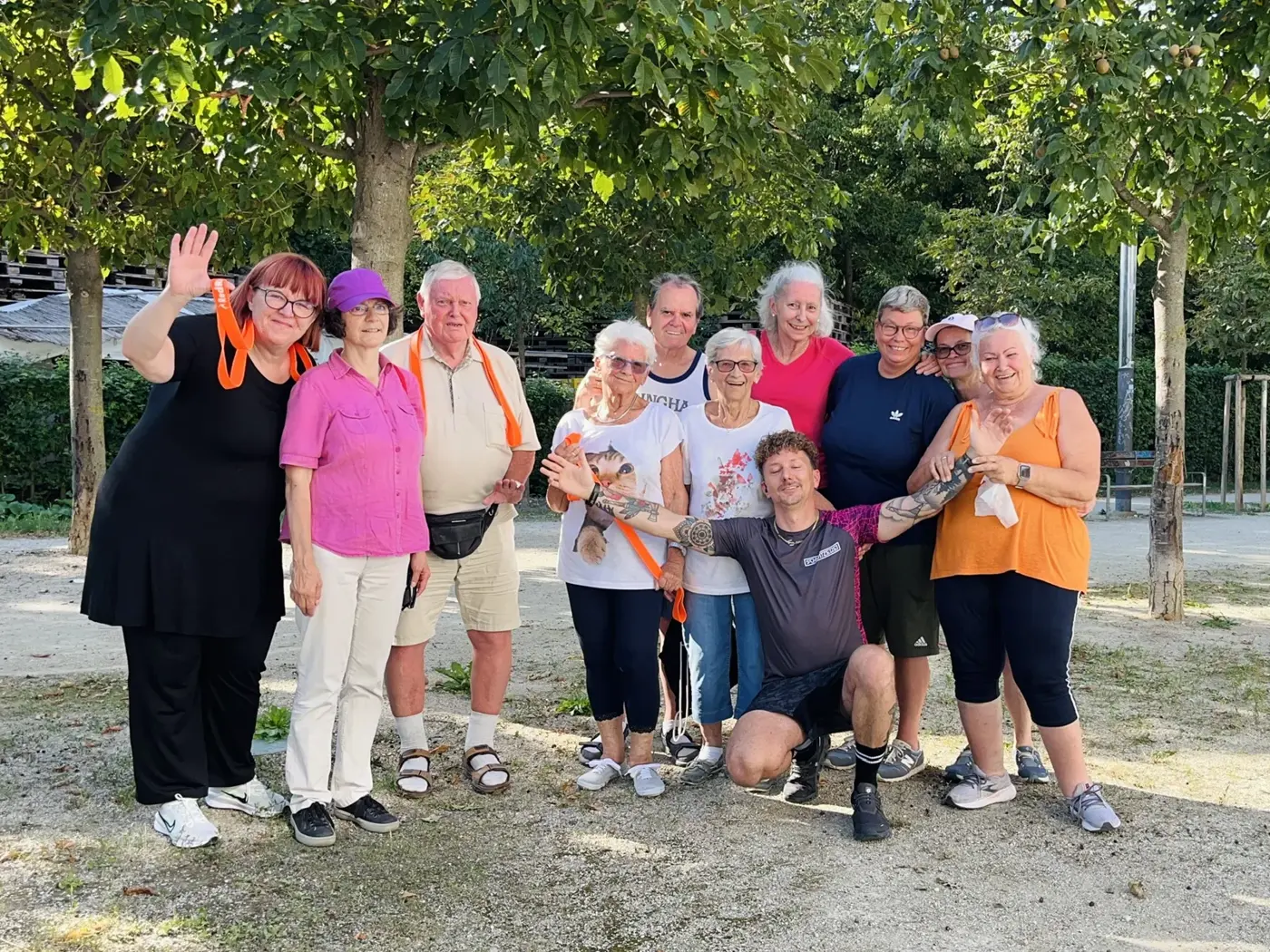 Die Gruppe winkt fröhlich in die Kamera. Trainer Manfred kniet vorne und breitet die Arme aus.